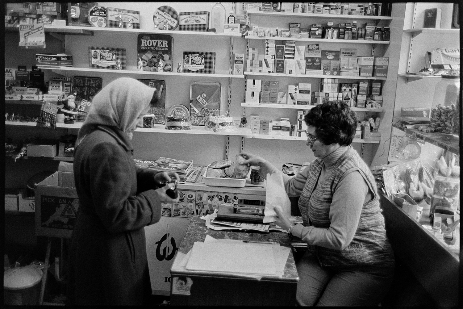 Women at till of grocers shop. 
[A woman serving a customer at the counter of the London House Stores in Fore Street, Dolton. Shelves with cigarettes, tins of biscuits and ornaments are visible in the shop.]
