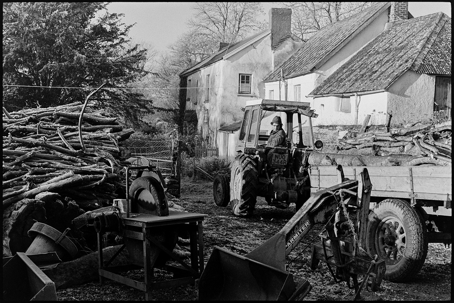 Farmer sorting logs in farmyard. 
[Stephen Squire driving a tractor pulling a trailer full of logs past the farmhouse at Lower Langham, Dolton. A woodpile and circular saw, covered with a tyre, can be seen in the farmyard.]
