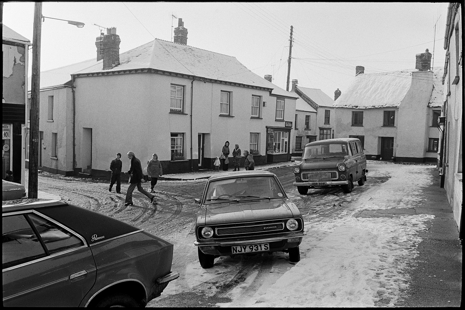 Street scene with cars in snow. 
[Cars and a van parked on a snow covered street in Winkleigh. People are walking along the street and the shop front of Winkleigh Post Office is visible in the background.]