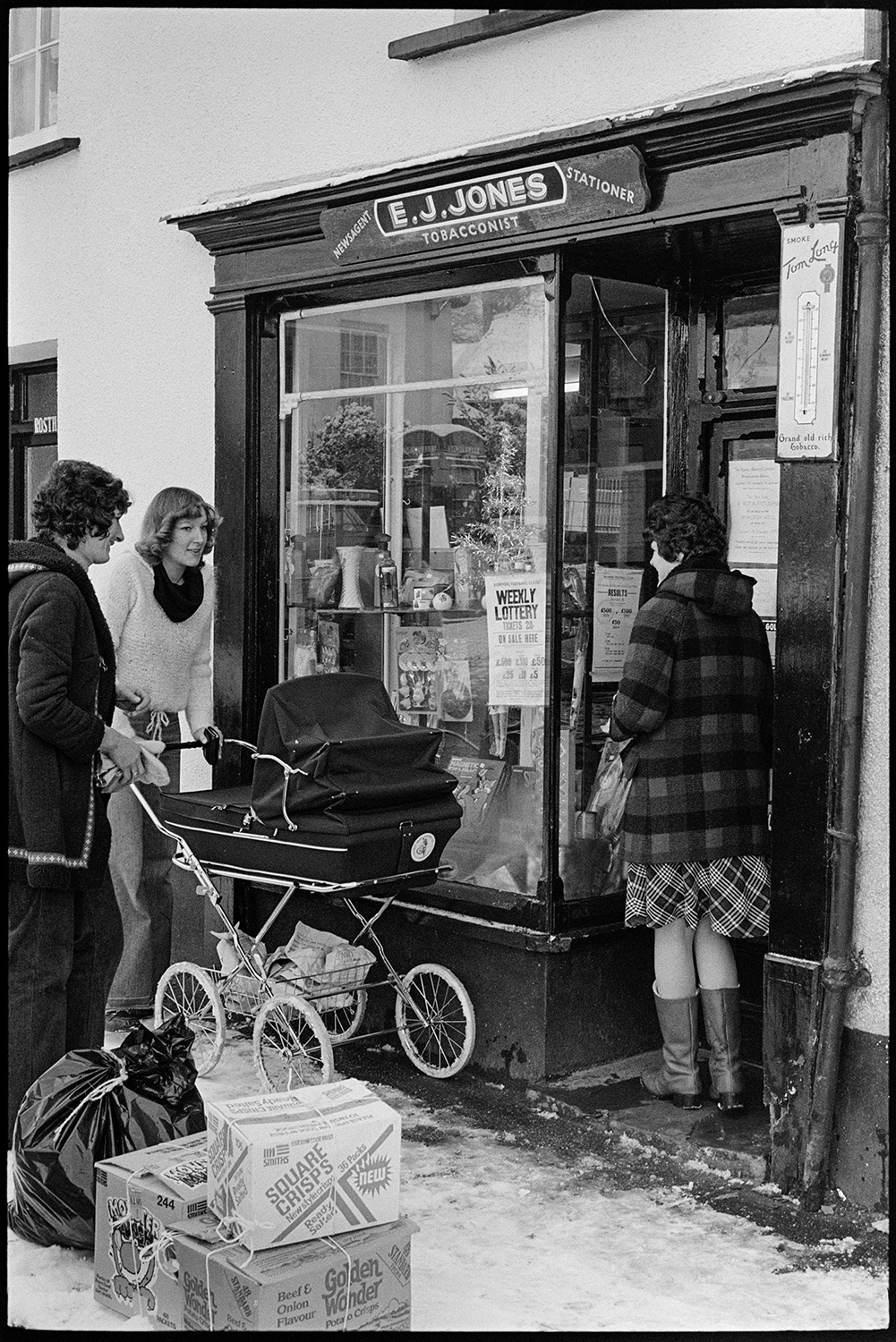 Shop front with women and pram. 
[People, including a woman with a pram outside the shop front of E J Jones, Tobacconist in Winkleigh. A bin bag and crisp boxes can be seen on the snow covered pavement outside the shop.]