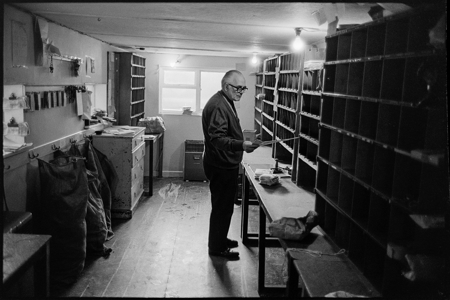 Sorting mail in sorting office. Franking mail. 
[Mr Butler sorting mail into pigeon holes in the sorting office at Winkleigh Post Office. Sacks of mail can be seen on the other side of the room.]