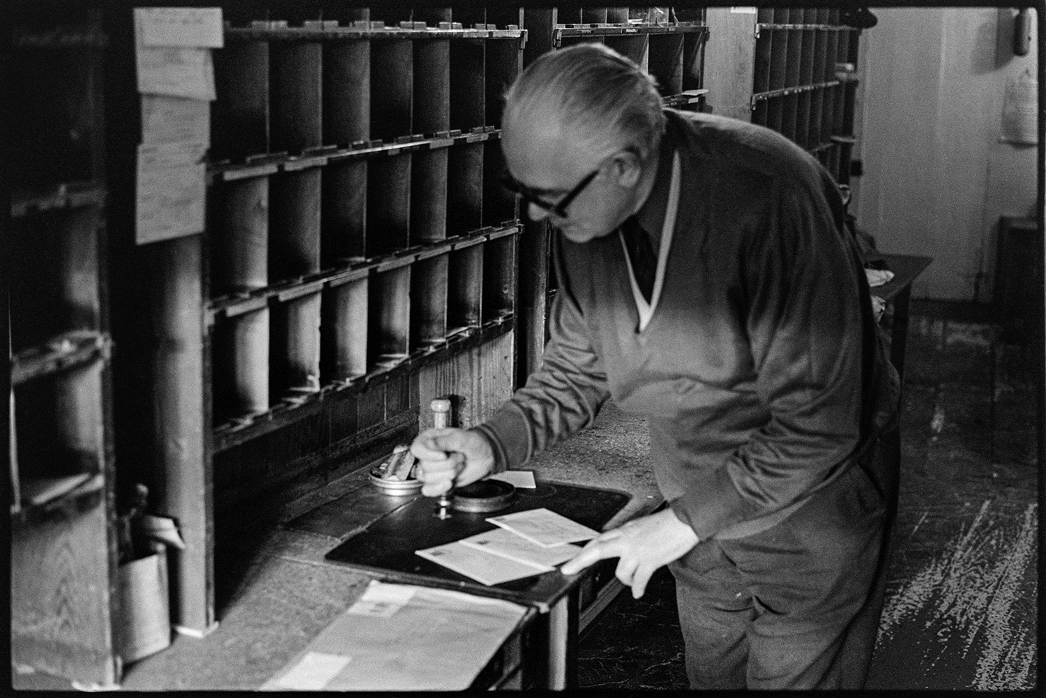 Sorting mail in sorting office. Franking mail. 
[Mr Butler franking mail using a stamp, by pigeon holes at Winkleigh Post Office.]