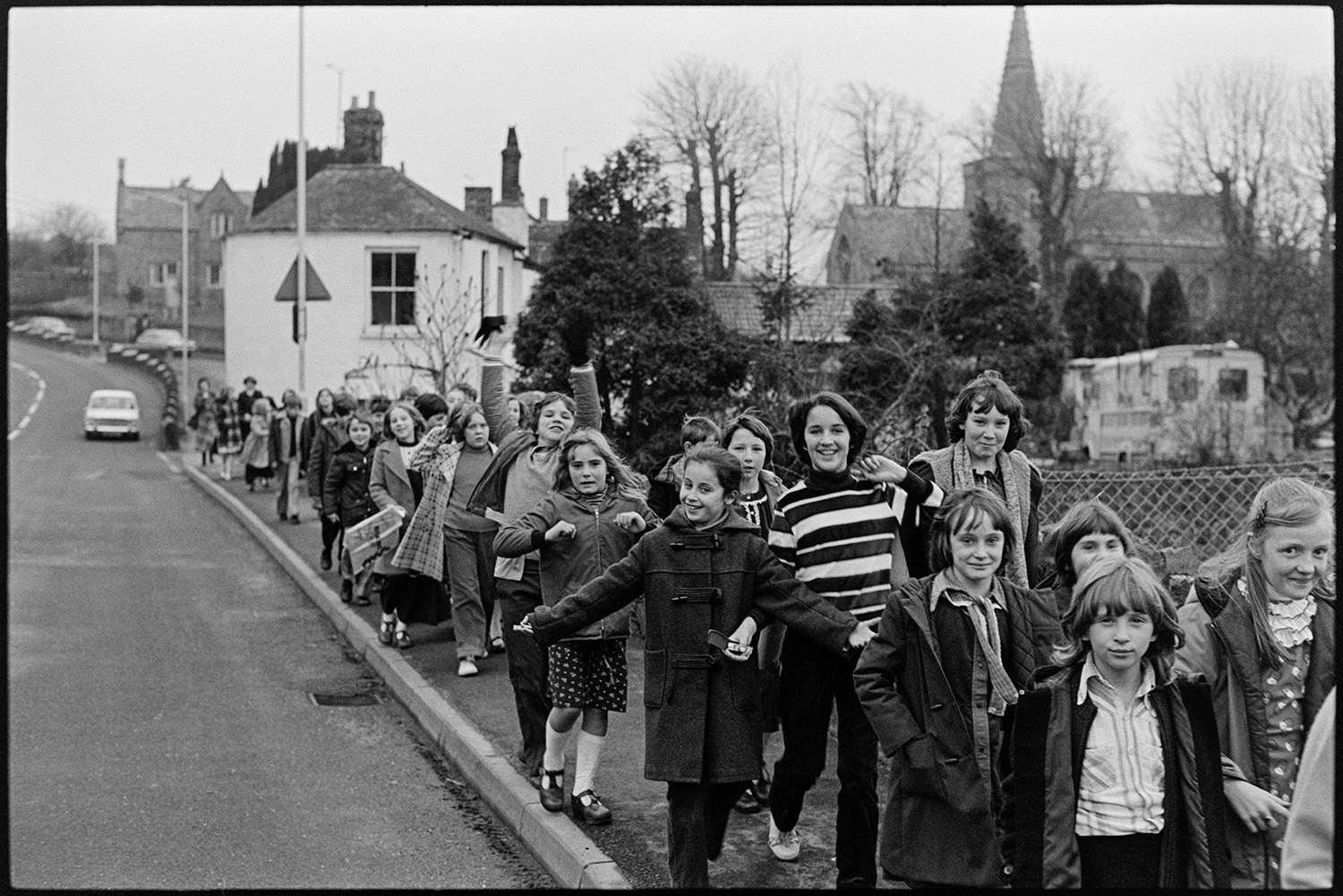 Crocodile of schoolchildren going through village. 
[A group of schoolchildren walking along a road through Bishops Tawton. Some of the children are waving. A church with a spire is visible in the background.]