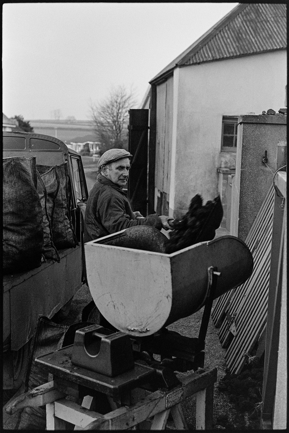 Coal merchant weighing, bagging and loading coal. 
[A man weighing coal using a weighing machine, outside a yard in Burrington. A truck with sacks of coal on it is visible beside him.]