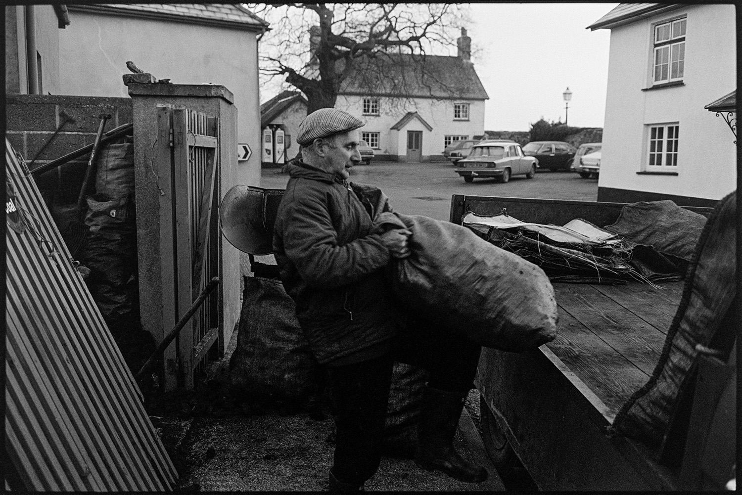 Coal merchant weighing, bagging and loading coal. 
[A man loading sacks of coal onto a truck in Burrington, Houses, parked cars, petrol pumps and the Burrington oak can be seen in the background.]