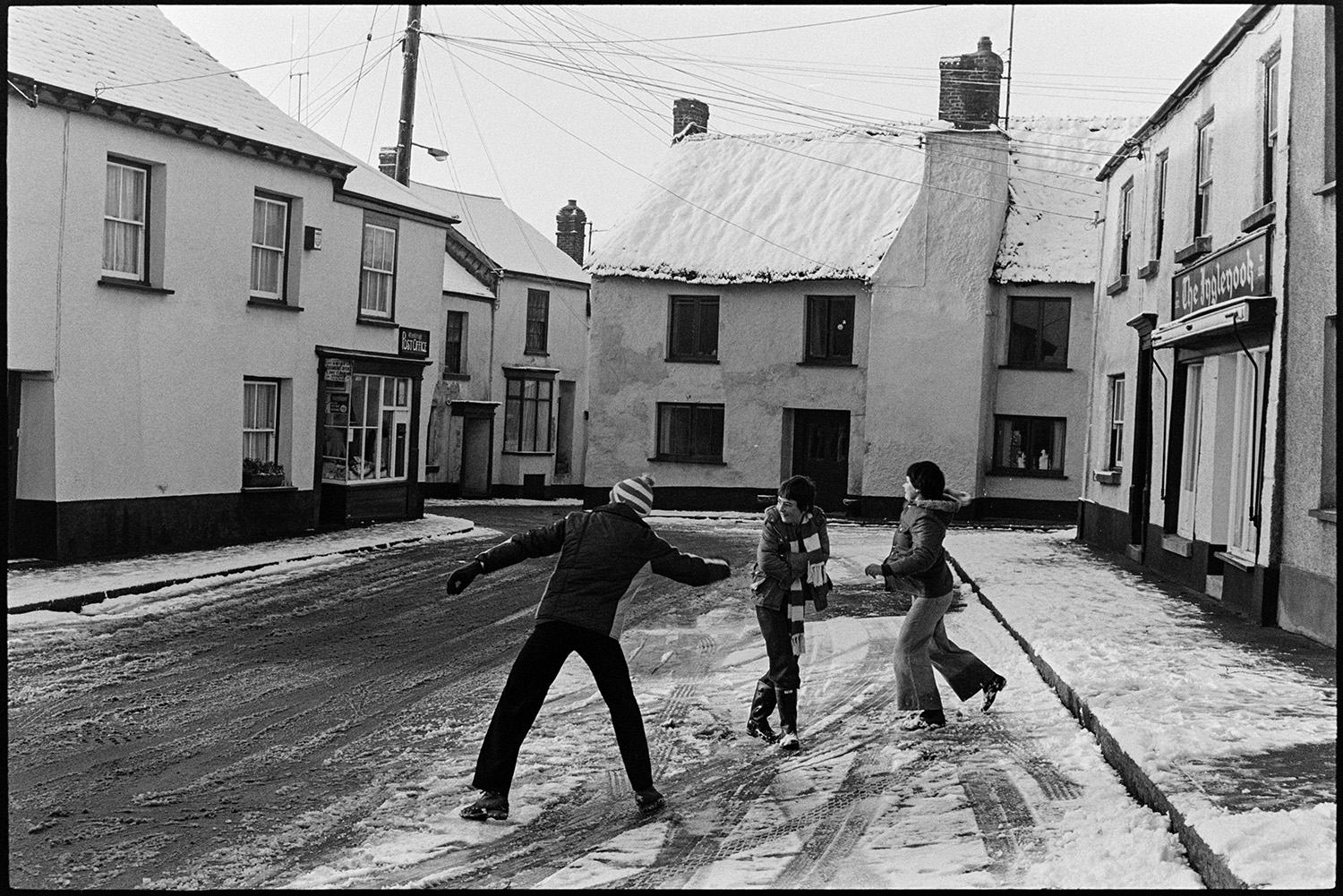 Children throwing snowballs in snowy village square. 
[Three children throwing snowballs in a snow covered street in Winkleigh. The Winkleigh Post Office is visible further down the street.]