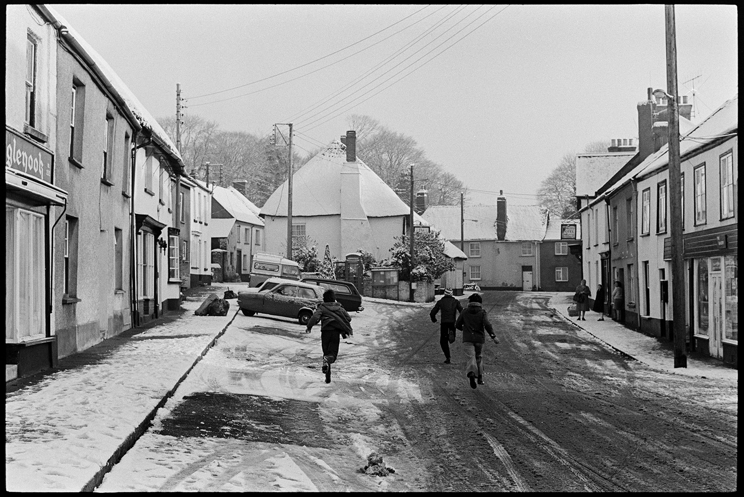 Children throwing snowballs in snowy village square. 
[Children running down a snow covered street in Winkleigh. Shop fronts and parked cars can be seen along the street.]