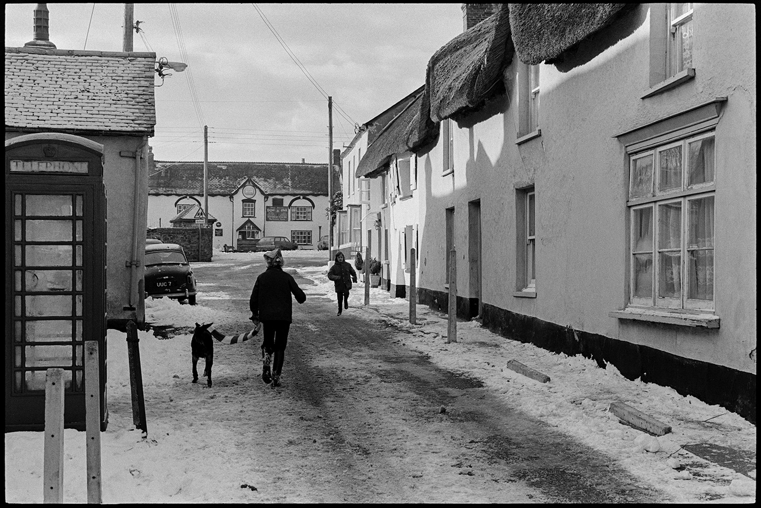 Snow, people shopping, chatting in snowy village, house with unusual roof. 
[People walking along Fore Street in Dolton, past thatched cottages and a telephone box. The street is covered with snow. The Royal Oak pub can be seen in the background.]