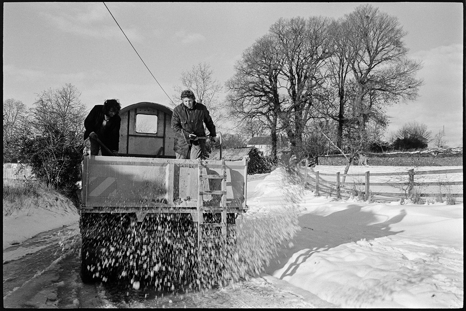 Snow, drift in road, men digging out gritting lorry and spreading from back, small barn in snow. 
[Men spreading grit from the back of a gritting lorry onto a road at Langham, Dolton, using shovels. Snow covered fields and trees are visible by the road.]