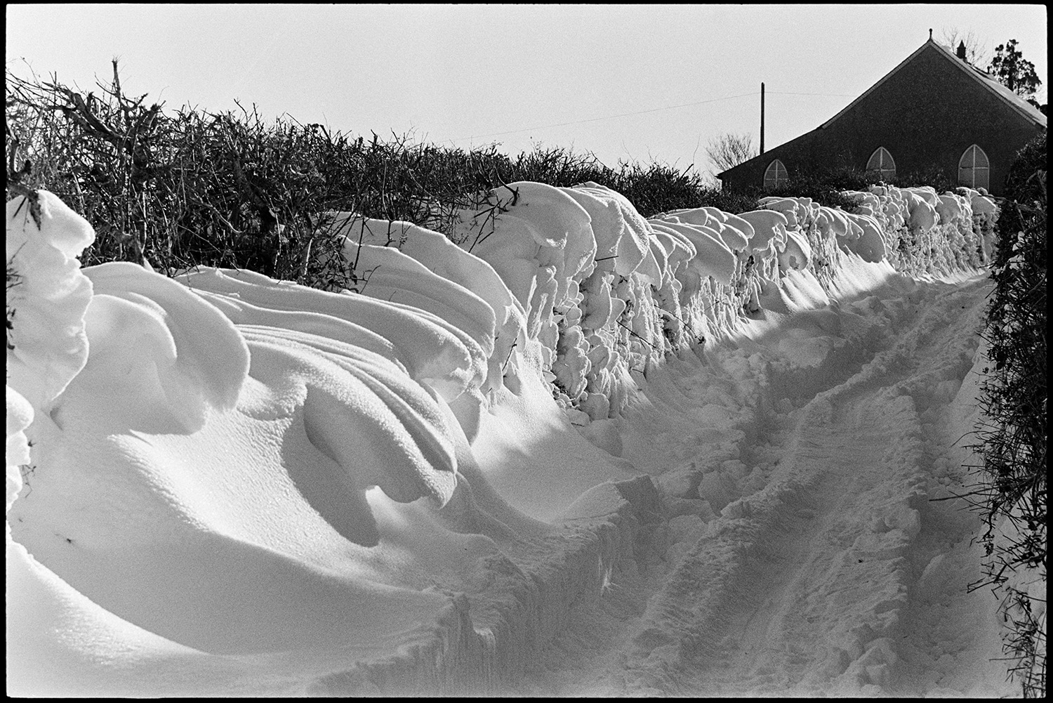Snow, drift in road, men digging out gritting lorry and spreading from back, small barn in snow. 
[A snowdrift against a hedge in a lane at Langham, Dolton. A building can be seen further along the lane.]