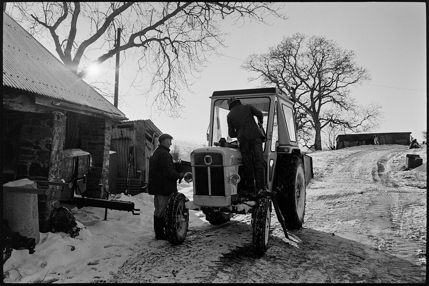 Snow, farmer trying to unfreeze tractor. 
[Two men trying to unfreeze a tractor in the snowy farmyard at Parsonage, Iddesleigh. Barns are visible in the background.]