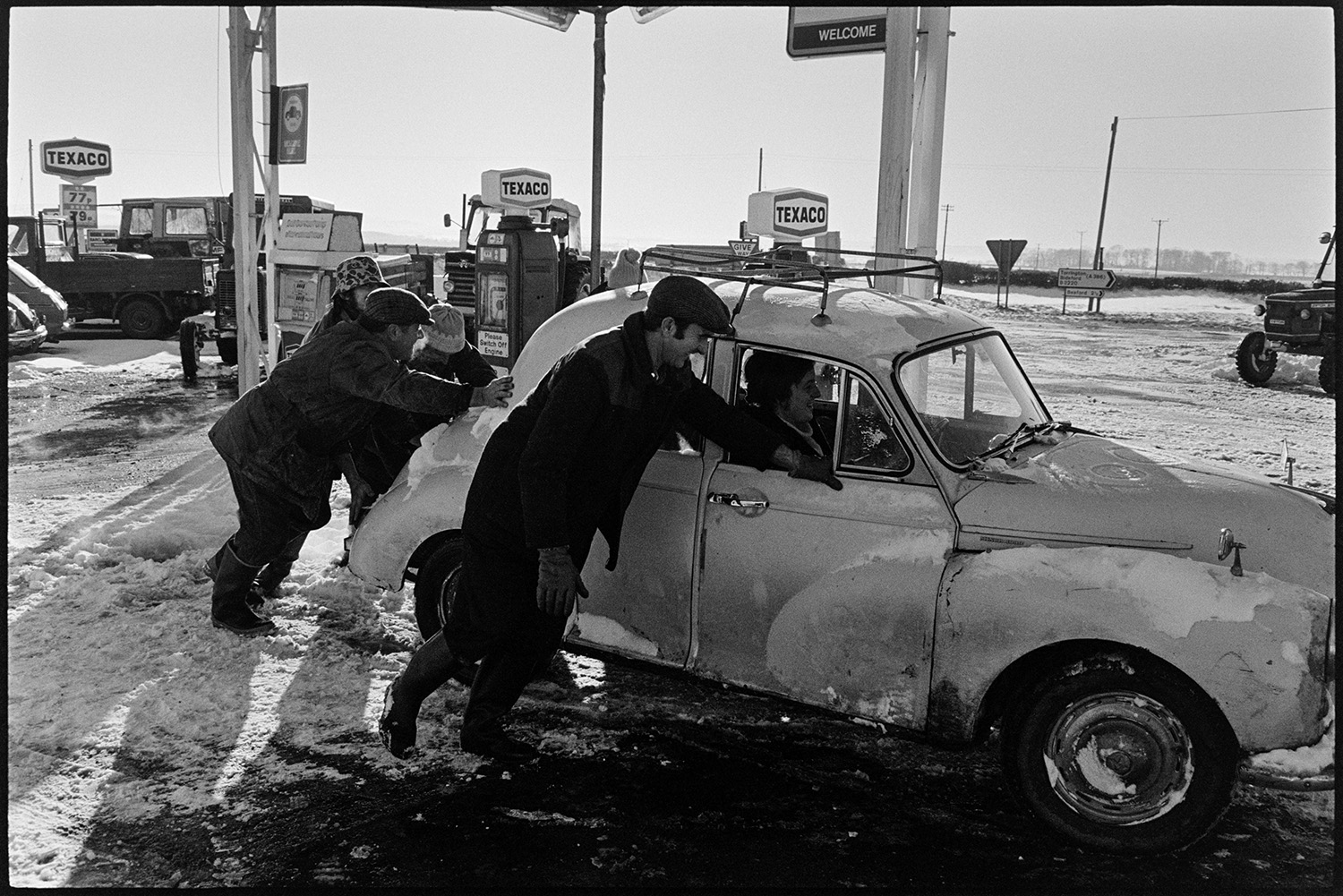 Snow, scenes garage forecourt trying to push start car. Bicycle. 
[A group of men trying to push start a car at Beacon Garage, Dolton. Texaco petrol pumps can be seen at the garage and the forecourt is covered with snow.]