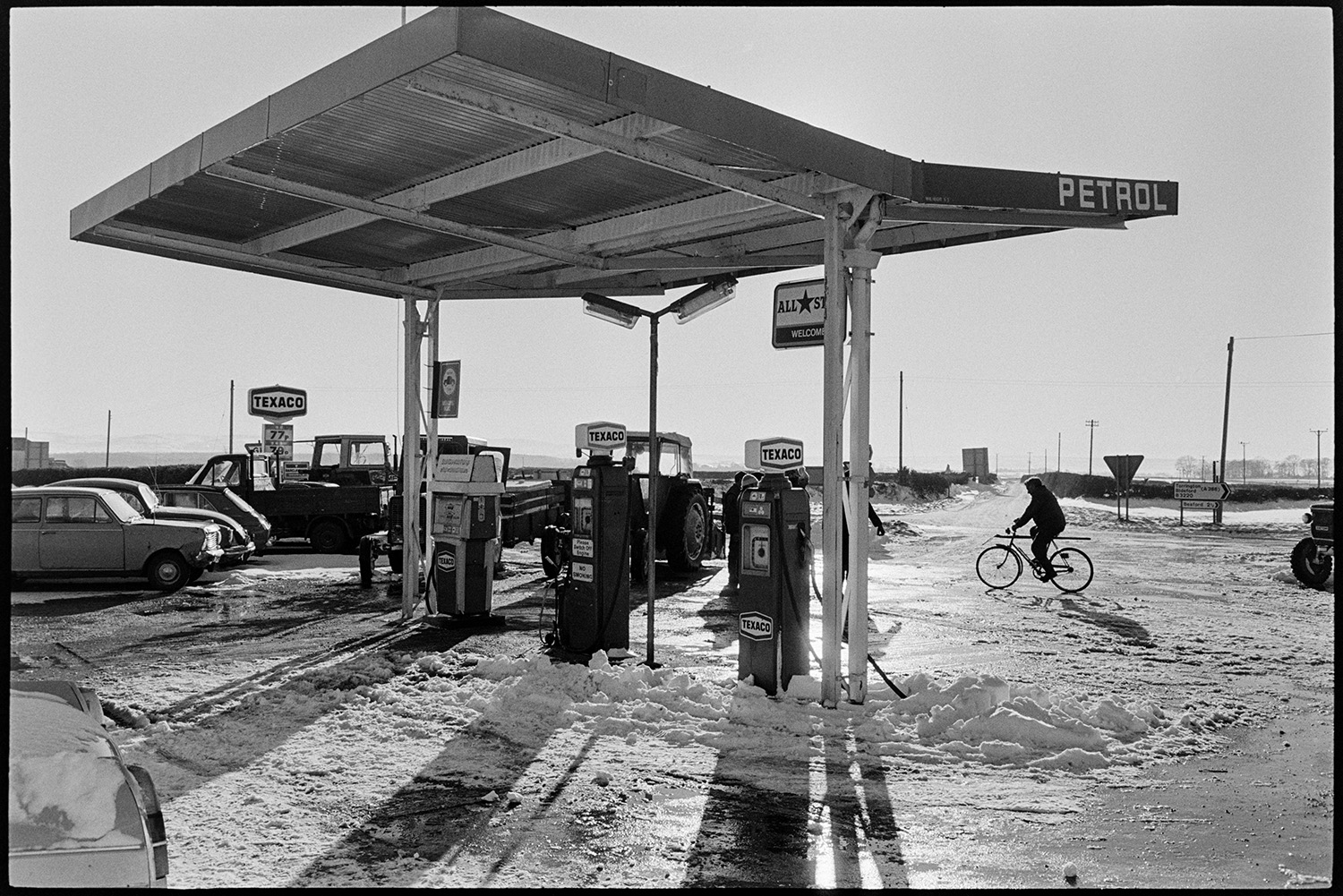 Snow, scenes garage forecourt trying to push start car. Bicycle. 
[A person cycling past Beacon Garage, Dolton, in the snow. Petrol pumps and parked cars and tractors can be seen at the garage.]