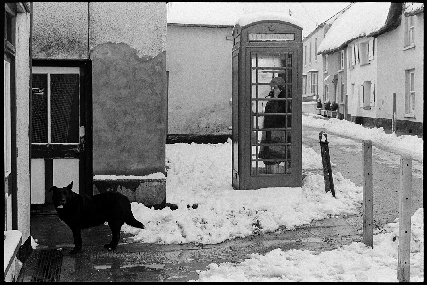 Snow, Street scenes with men spreading grit on road, woman telephoning from kiosk. 
[A woman using the telephone box in Fore Street, Dolton. The street and thatched cottage outside are covered with snow and a dog is waiting outside Dolton Post Office.]