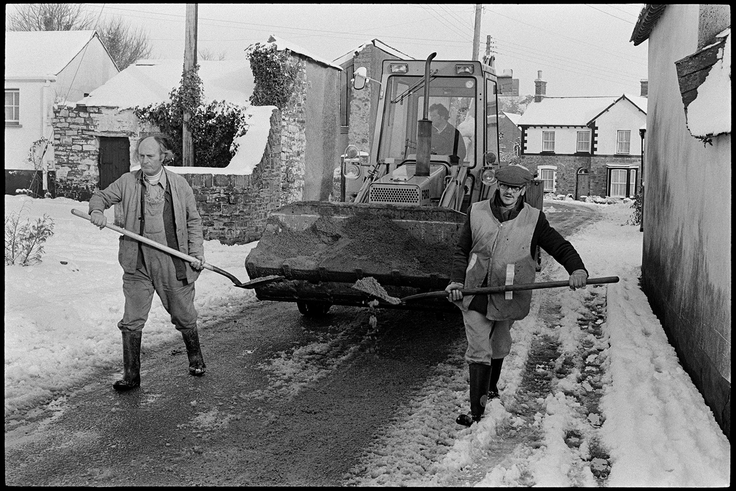 Snow, Street scenes with men spreading grit on road, woman telephoning from kiosk. 
[Men spreading grit on a snowy road in Dolton, using shovels. A tractor is following them with a load of grit in its bucket.]