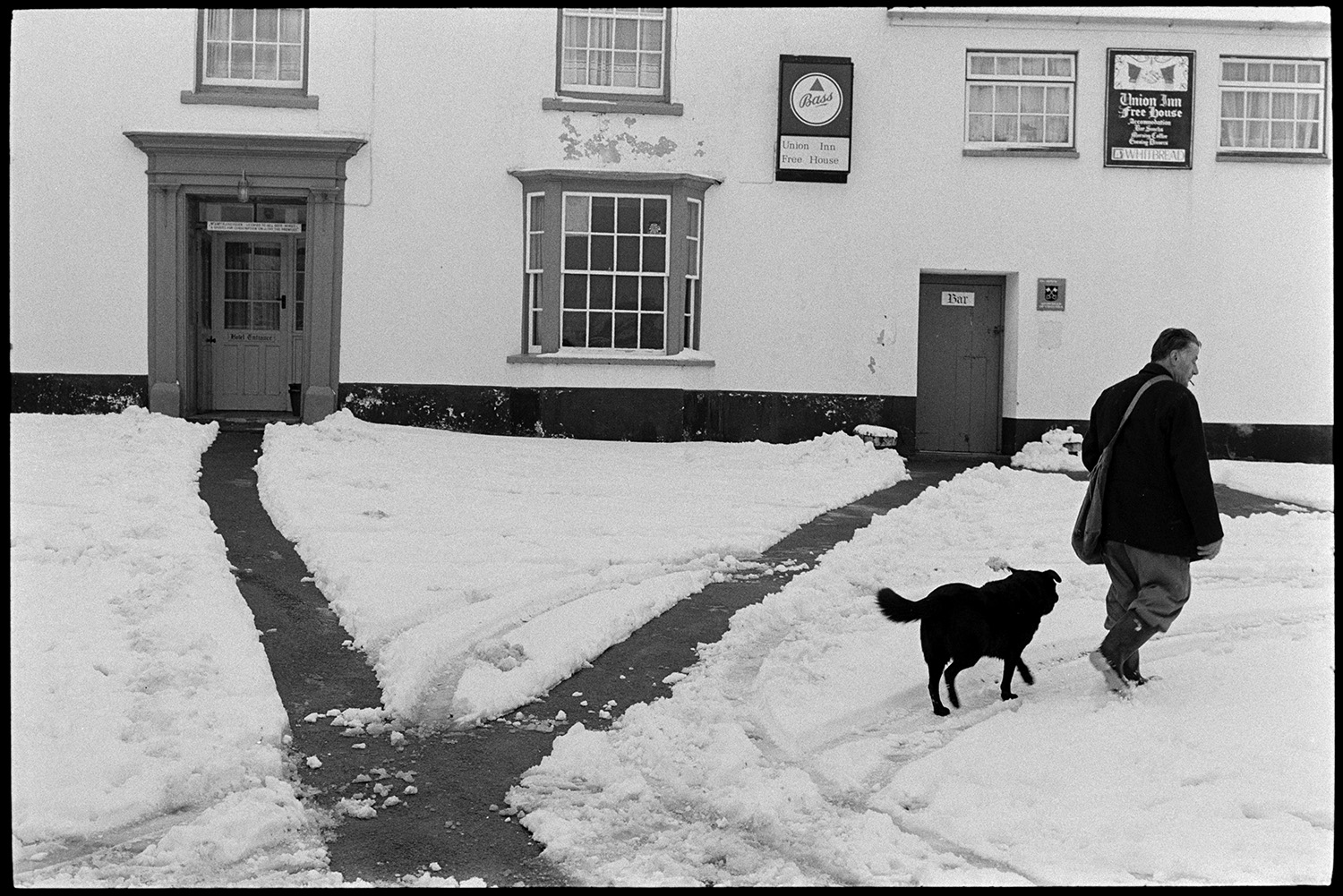Snow, Street scenes postman and dog, children throwing snowballs, people chatting. 
[Harold Nott, also known as Joe Bec, a postman, walking through snow outside the Union Inn pub in Dolton. He is accompanied by a dog. Two pathways have been cleared through the snow to the pub doors.]