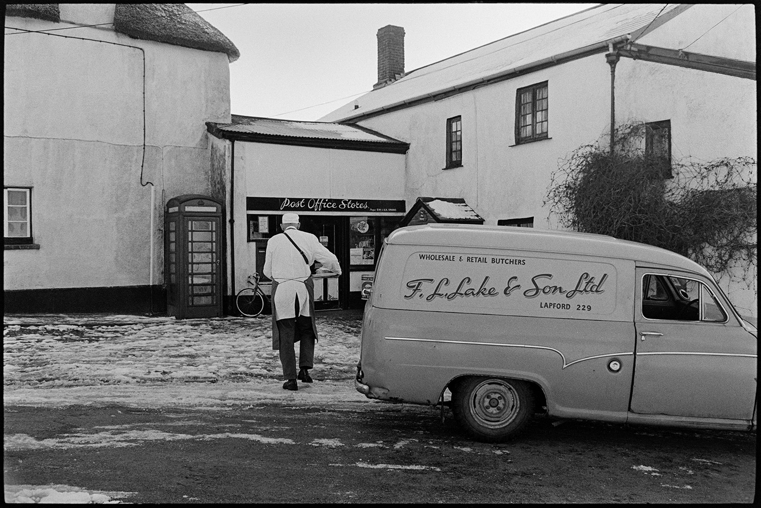 Snow, Butcher delivering meat to Post Office from van. 
[A butcher from F L Lake & Sons Ltd delivering meat to the Post Office in Coldridge. Snow is on the street and a telephone box is next to the Post Office.]