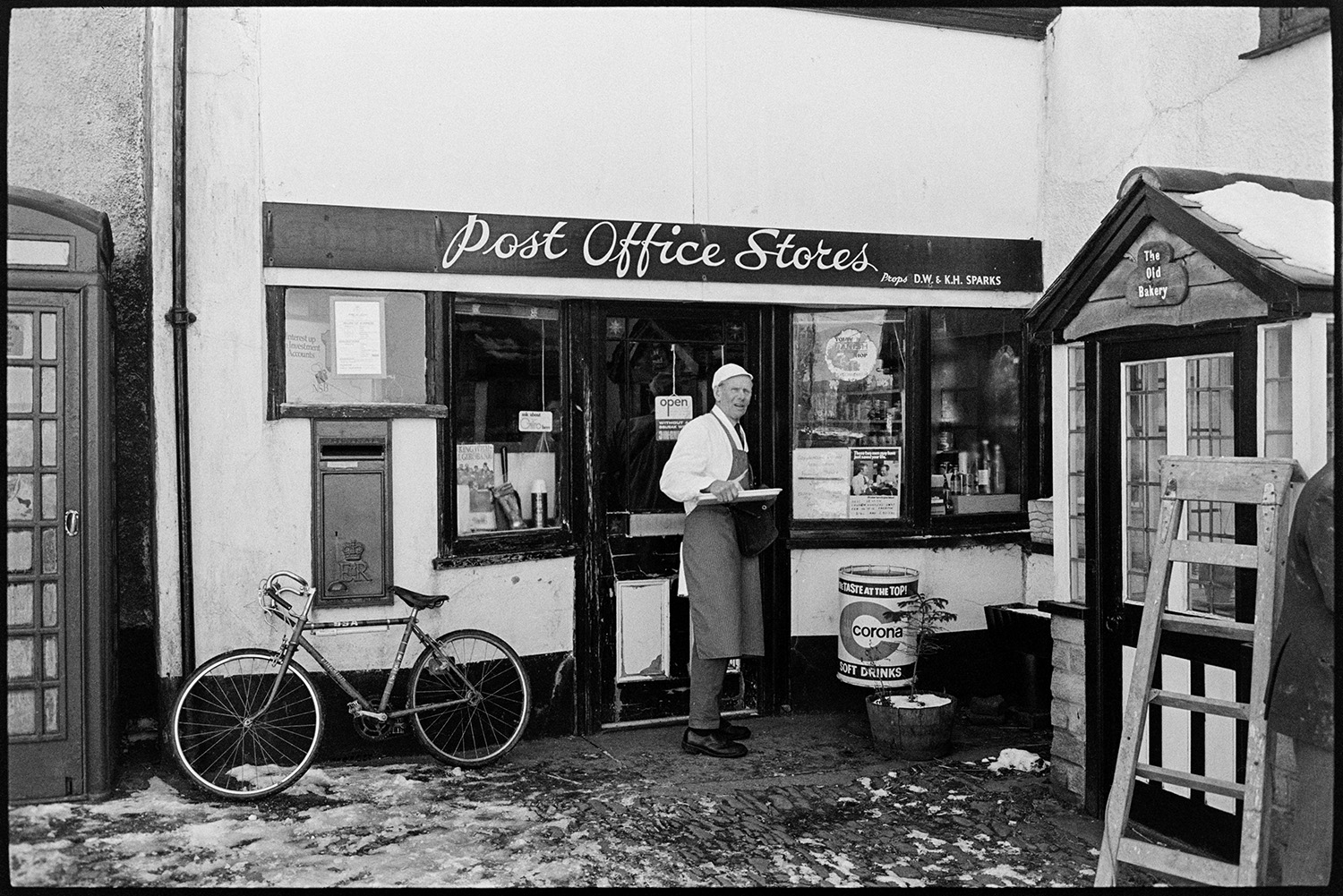 Snow, Butcher delivering meat to Post Office from van. 
[A butcher from F L Lake & Sons Ltd outside the Post Office in Coldridge, after delivering meat to the shop. A bicycle is lent against the shop front, under the post box , and a telephone box is next to the store. Snow can also be seen on the cobbles in front of the shop.]