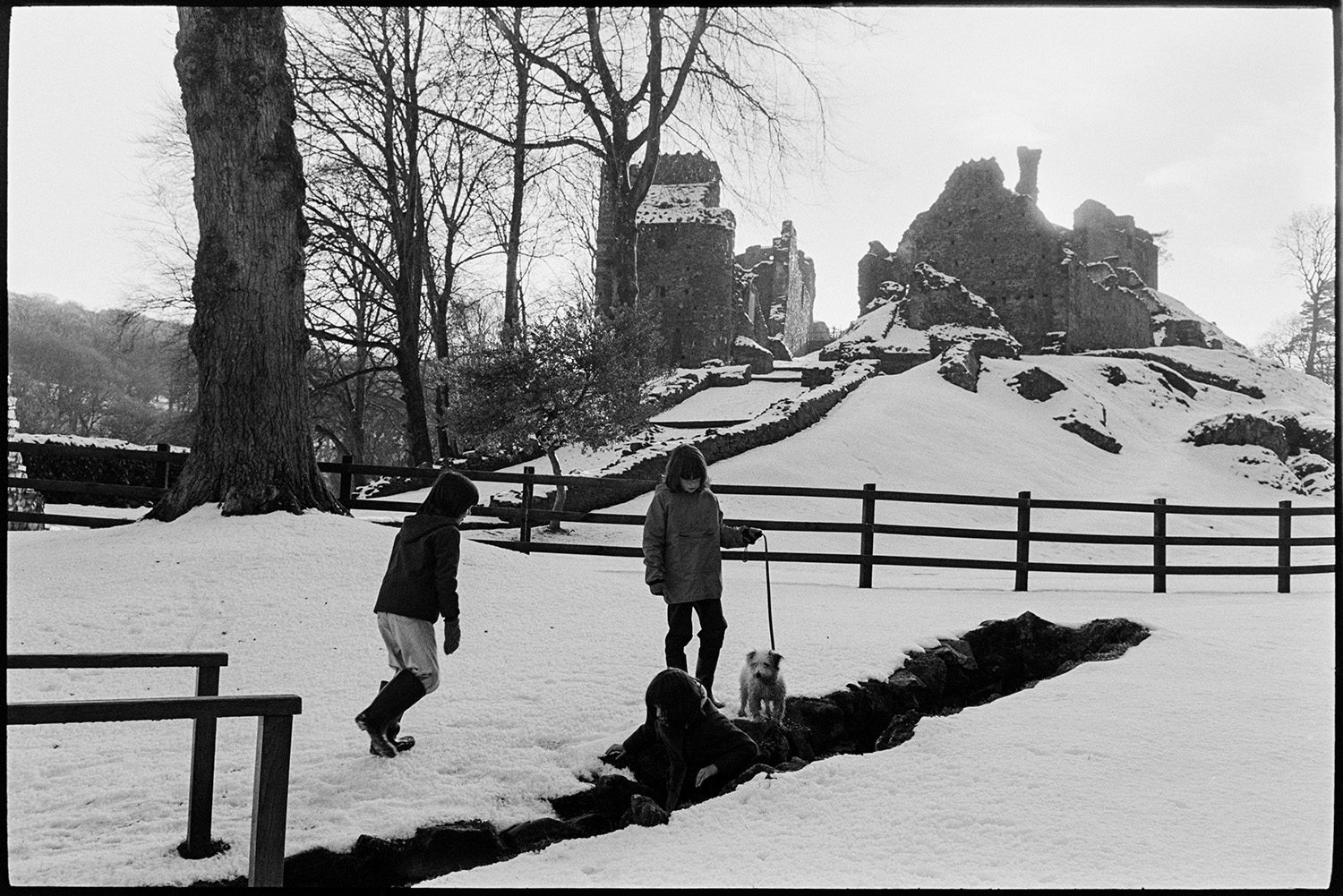 Snow, children with dog in front of snow covered castle. 
[Children and a dog playing in a trench in the snow by the ruins of Okehampton Castle.]