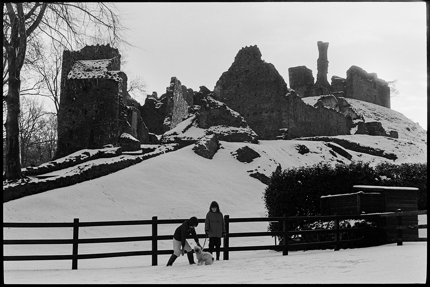 Snow, children with dog in front of snow covered castle. 
[Children playing with a dog in the snow by the ruins of Okehampton Castle.]