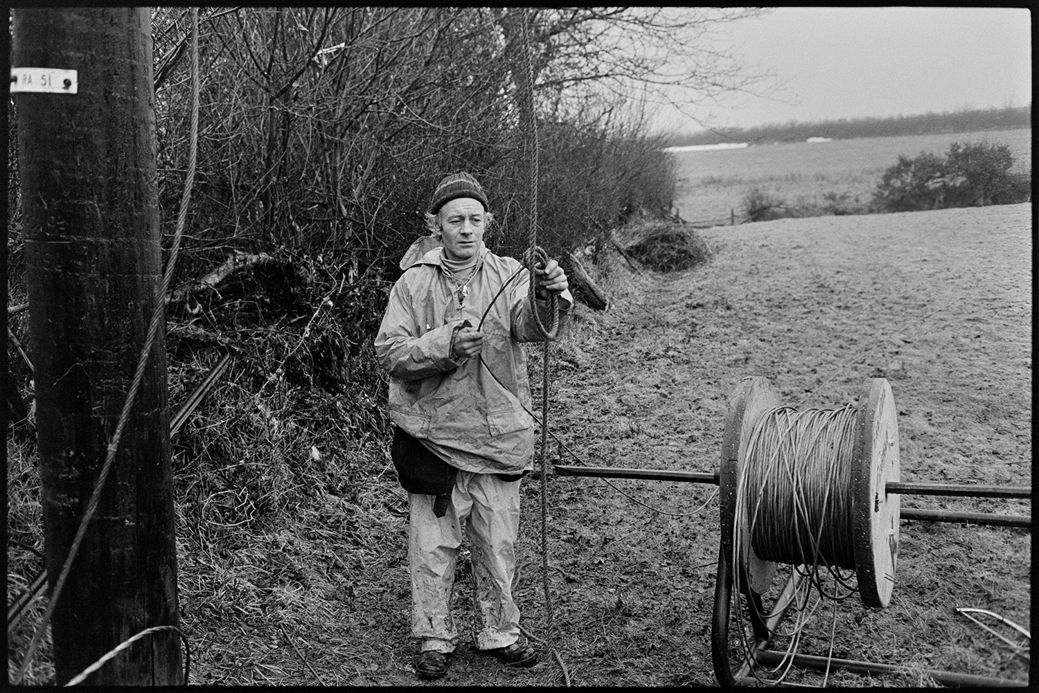 Men replacing electricity power lines, Land Rover and drums of cable. 
[A man from SWEB replacing electricity power cables at Dolton. He is stood in a field by a utility pole, next to a drum with a large reel of cable.]