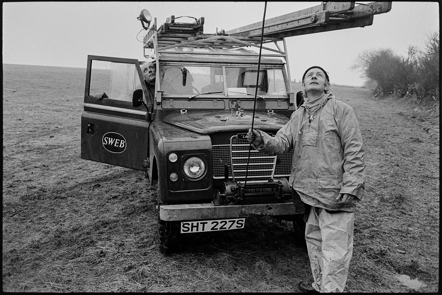 Men replacing electricity power lines, Land Rover and drums of cable. 
[Men from SWEB replacing electricity power cables at Dolton. They are in a field with a Land Rover. One of the men is holding a cable attached to the Land Rover.]