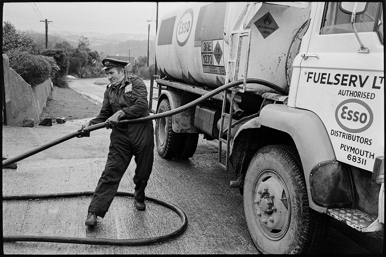 Man connecting hose from fuel oil tanker lorry to house storage. 
[A man holding a hose from an Esso fuel tanker, filling up the oil tank of a house in West lane, Dolton.]