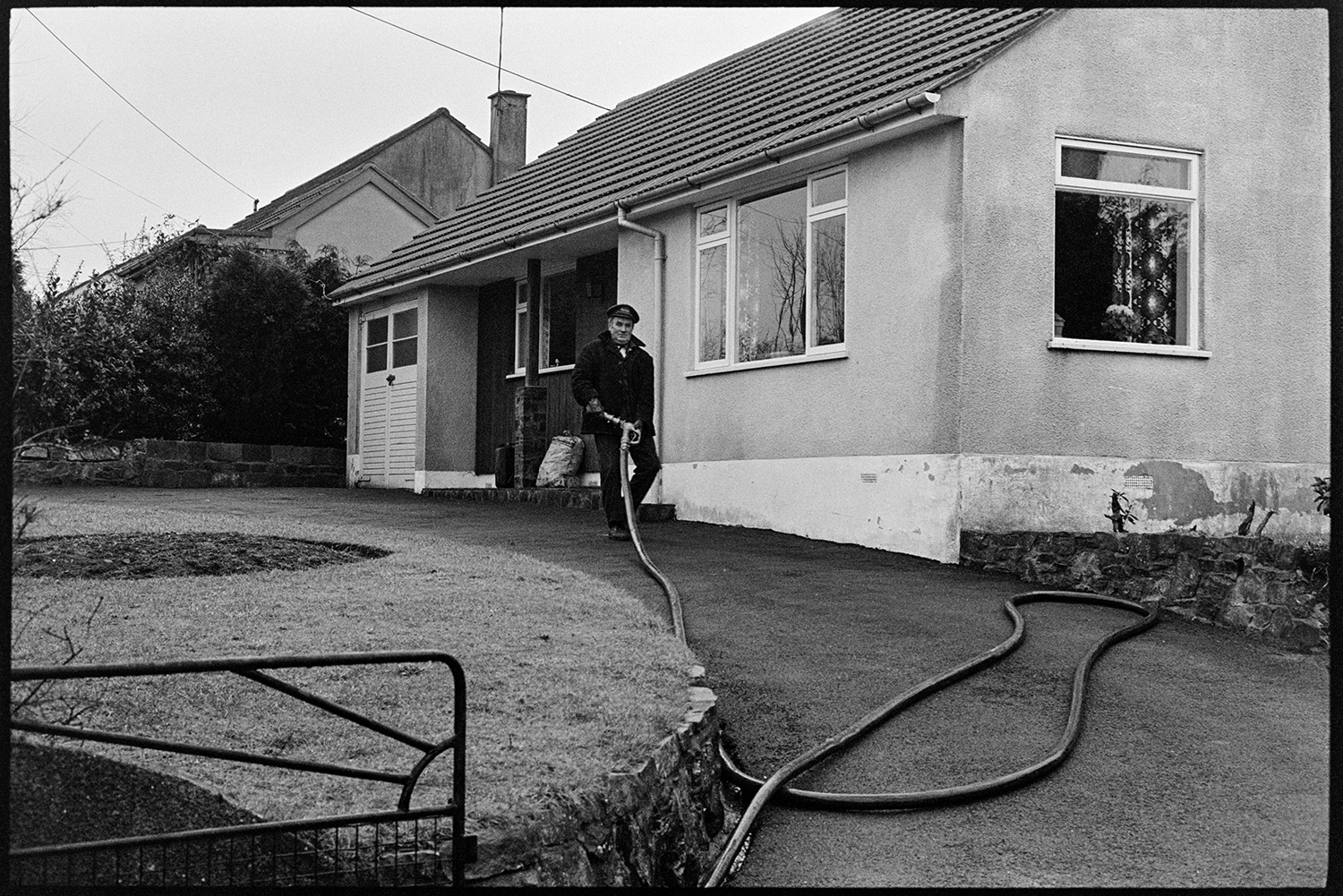 Man connecting hose from fuel oil tanker lorry to house storage. 
[A man running a hose from an Esso fuel tanker to a bungalow in West Lane, Dolton, to fill up the bungalow's oil tank.]