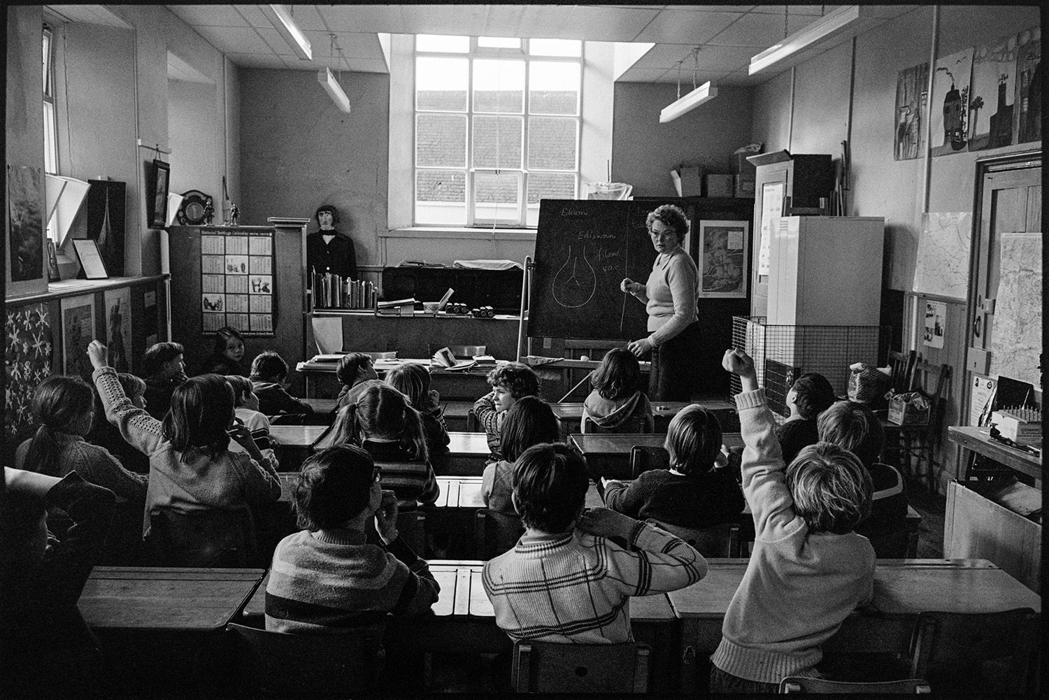 Primary school classroom with children listening to radio schools broadcast and working. 
[A woman teaching a classroom of children at Burrington Primary School. Two of the children have their hands up to answer a question. The teacher has drawn a bulb on the blackboard.]