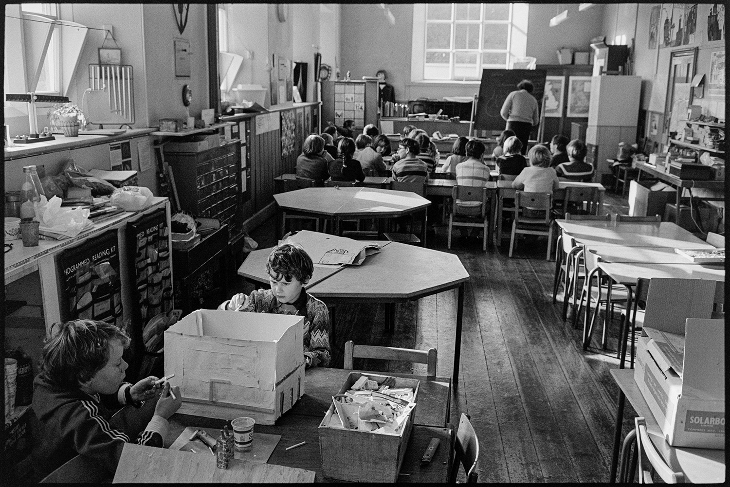 Primary school classroom with children listening to radio schools broadcast and working. 
[A teacher writing on a blackboard in a classroom at Burrington Primary School. Children are sitting and watching. Two children in the foreground are making a house from a cardboard box.]