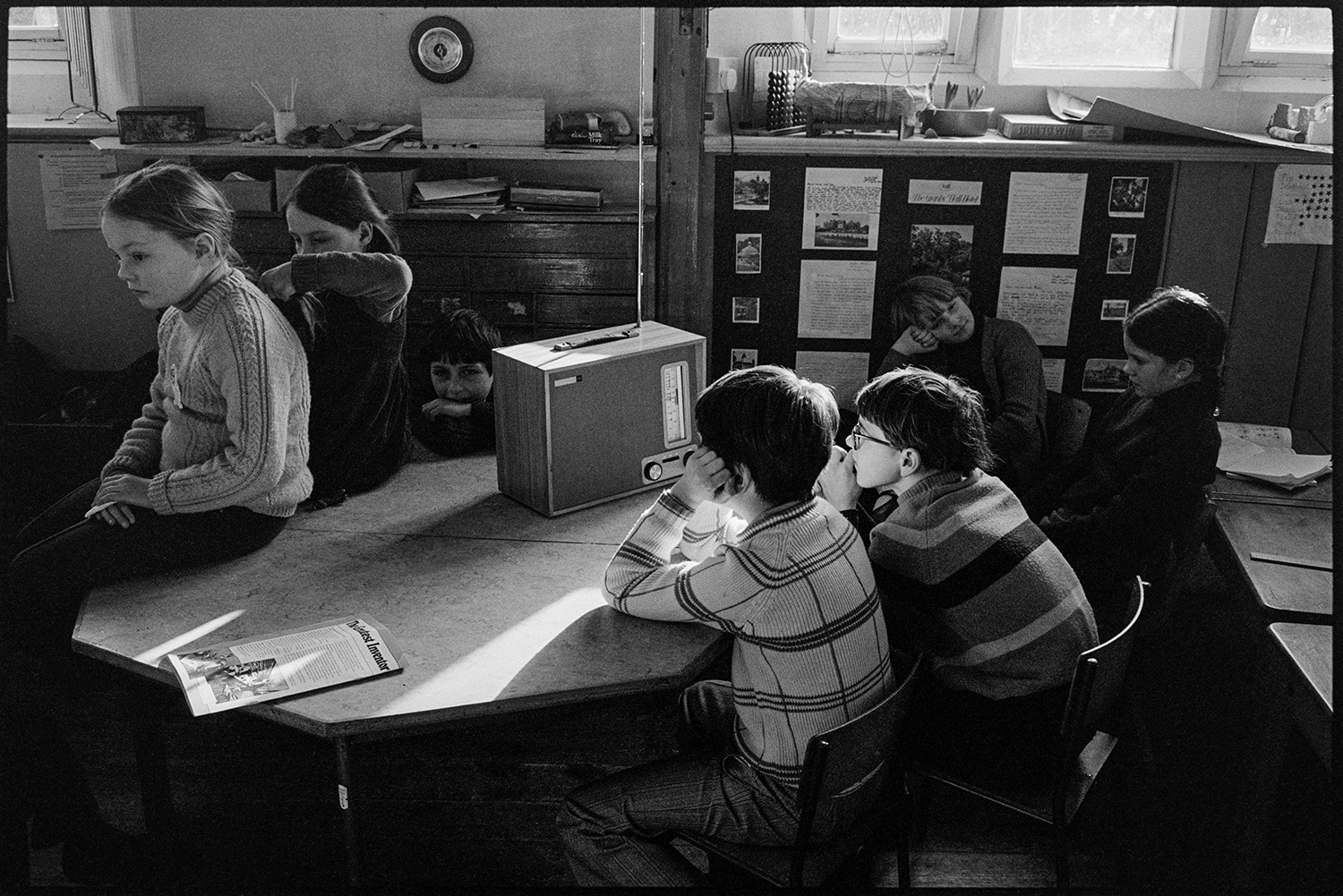 Primary school classroom with children listening to radio schools broadcast and working. 
[Children sat around, and on, a table in a classroom at Burrington Primary School. They are listening to a schools broadcast from the radio on the table.]