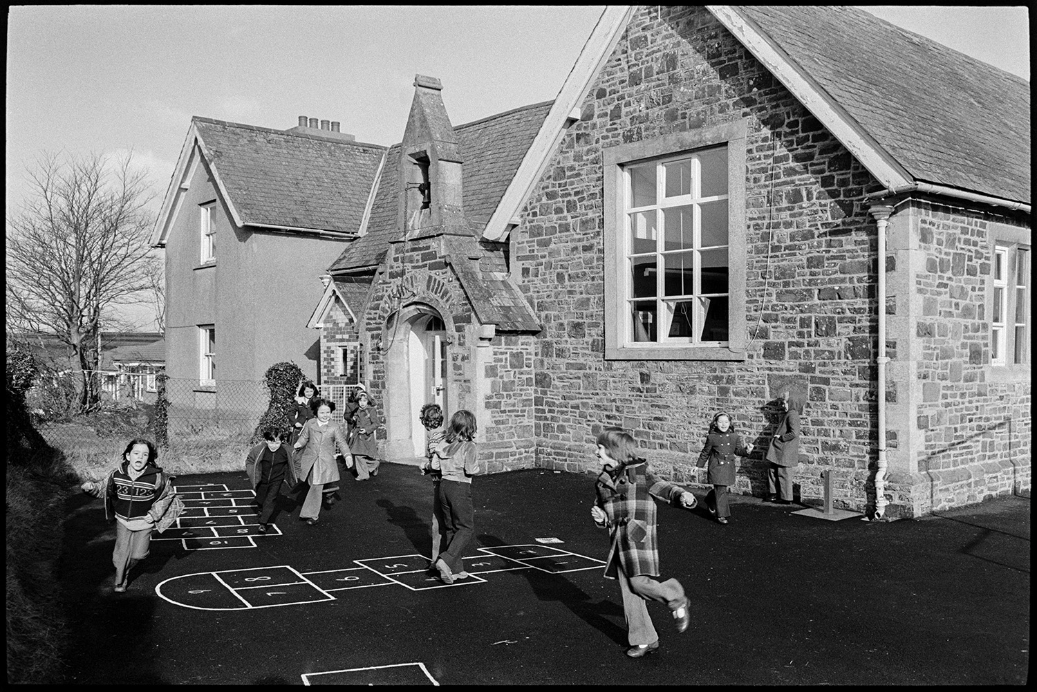 School children playing in playground and on village green, the kitchen. 
[Schoolchildren playing and running around the playground at Burrington Primary School. Hopscotch are painted on the playground floor.]