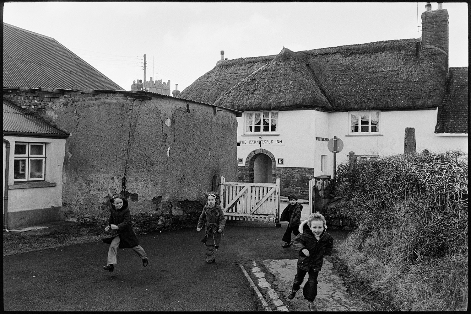 School children playing in playground and on village green, the kitchen. 
[Schoolchildren walking in through the school gate to Burrington Primary School. The thatched Barnstaple Inn can be seen in the background.]
