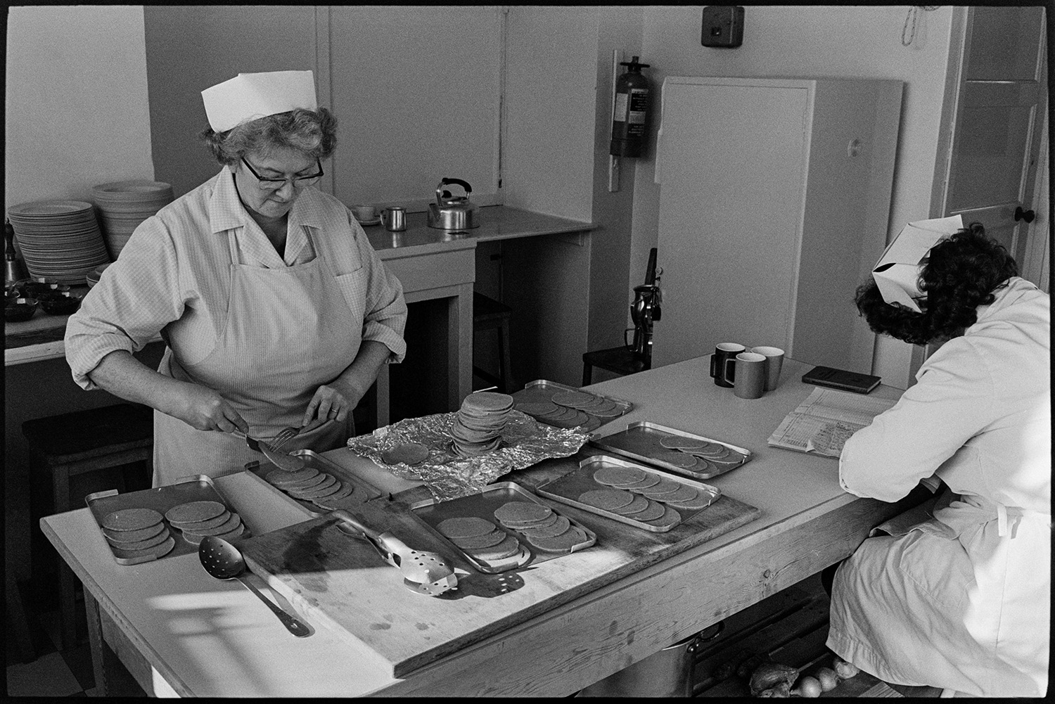 School dinner ladies doing accounts and preparing food in the kitchen. 
[School dinner ladies in the school canteen at Burrington Primary School. One is preparing food and the other is writing up the accounts in a ledger.]