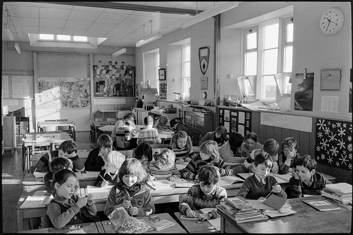 Primary school classroom with headmistress teaching, views of full classroom. 
[Schoolchildren working in a classroom at Burrington Primary School. Their school work is displayed on the walls of the classroom.]