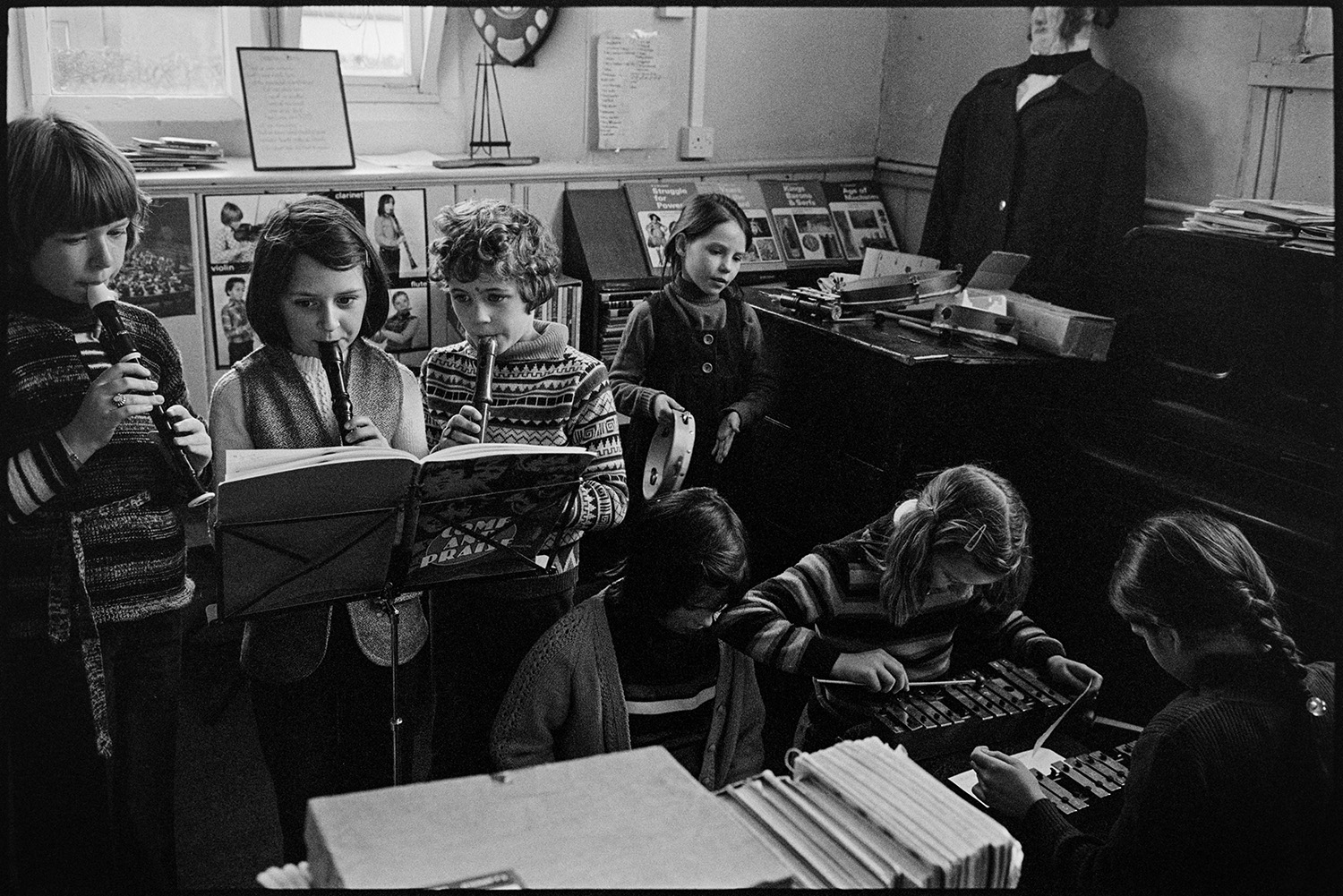 School children playing music and having work corrected. 
[Schoolchildren playing musical instruments, including recorders, a tambourine and xylophones at in a classroom at Burrington Primary School.]