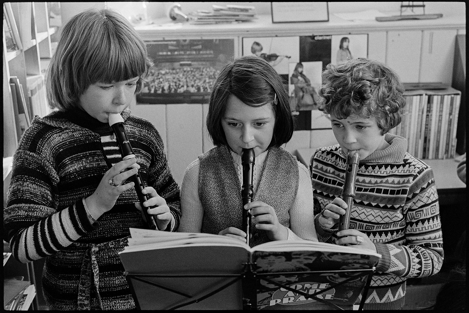 School children playing music and having work corrected. 
[Three schoolchildren playing recorders at Burrington Primary School. They are playing from a music book on a music stand.]