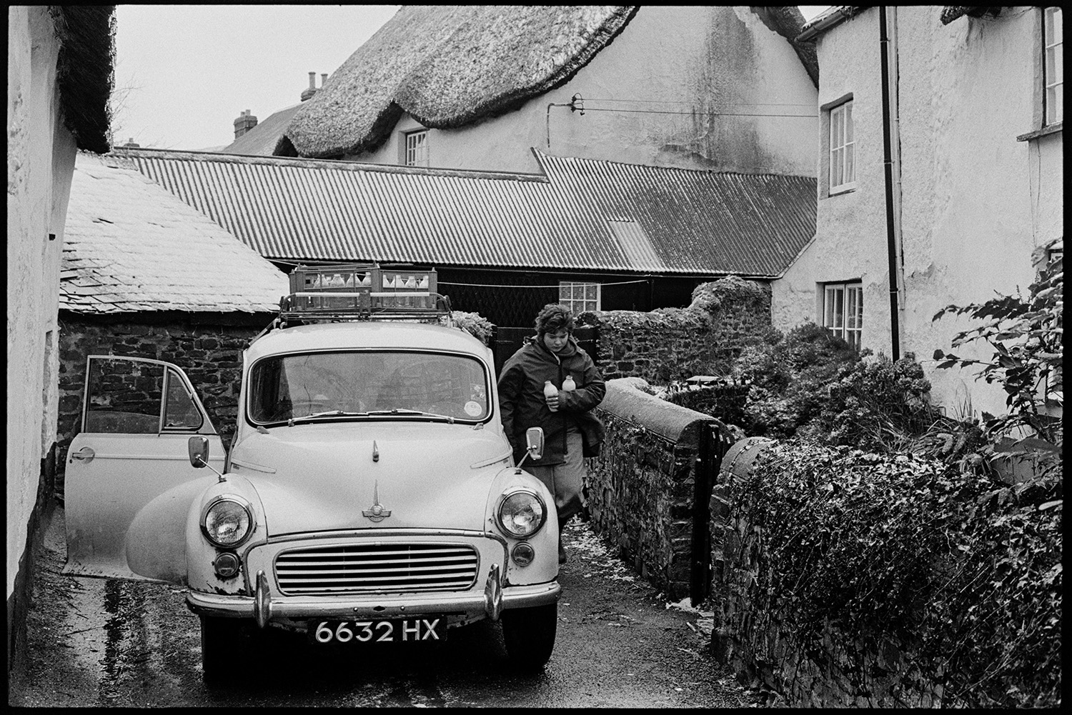 Woman delivering milk round village from Morris Minor car. 
[Diane Hiscock delivering milk around Dolton in her Morris Minor car. She is taking two milk bottles to a house and crates of milk are stacked on top of the car.]