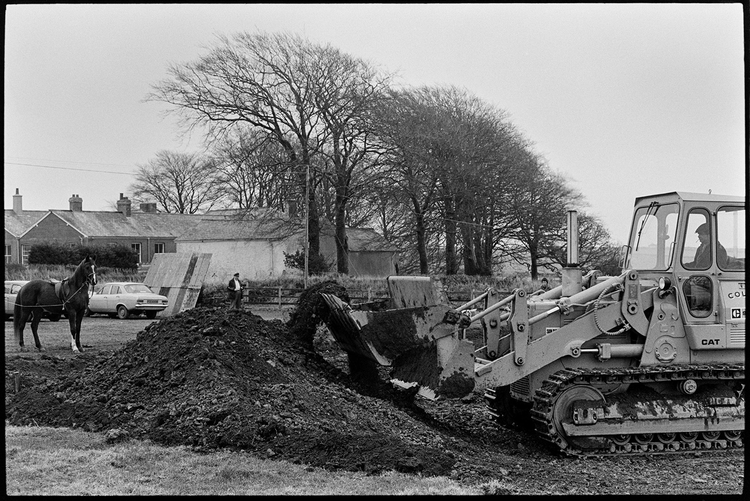 Man training horse getting it to tolerate bulldozer noise. New village hall foundations. 
[A bulldozer digging foundations for the new village hall in Beaford. A horse is visible in the background which is being trained by Henry Bright to get used to bulldozer noise.]