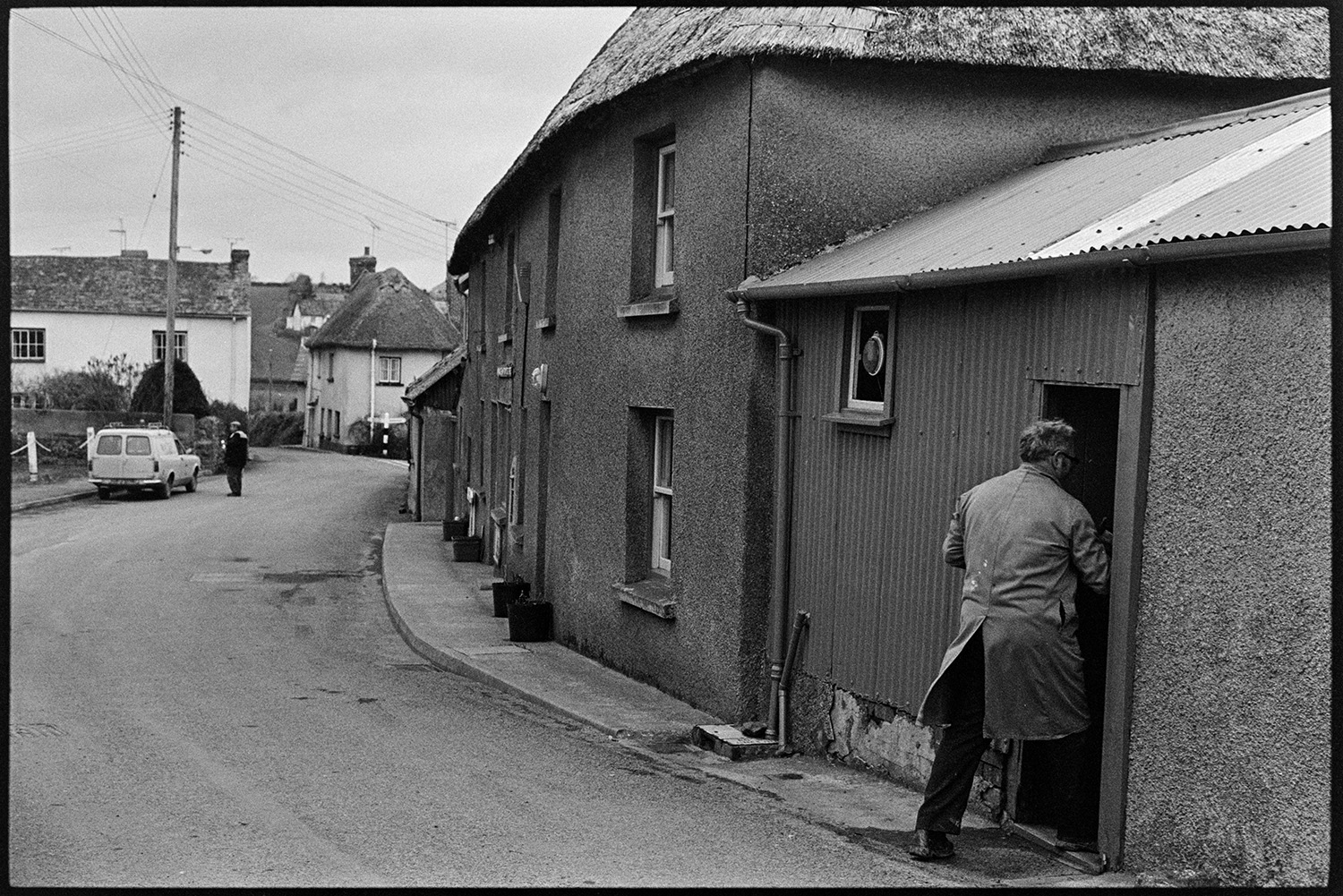Street scene with man going into door of workshop. 
[A man going into a workshop with a corrugated iron roof and attached to a thatched cottage, in a street in Monkokehampton.]