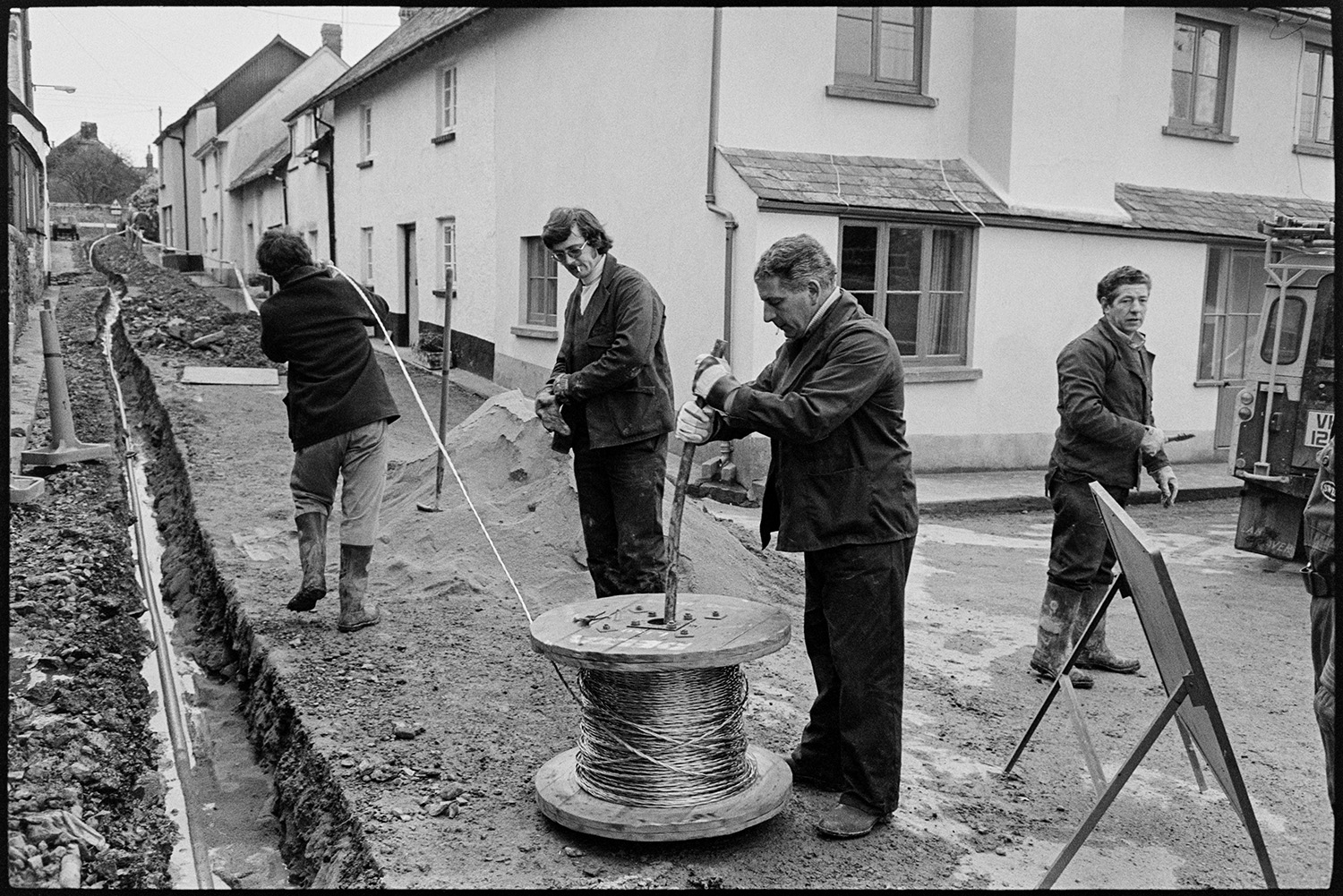 Men laying main electricity cable in village street, cable drum on trailer, drills etc. 
[Men unravelling an electricity cable from a cable drum in Church Street, Dolton, before laying it in a trench dug through the street.]