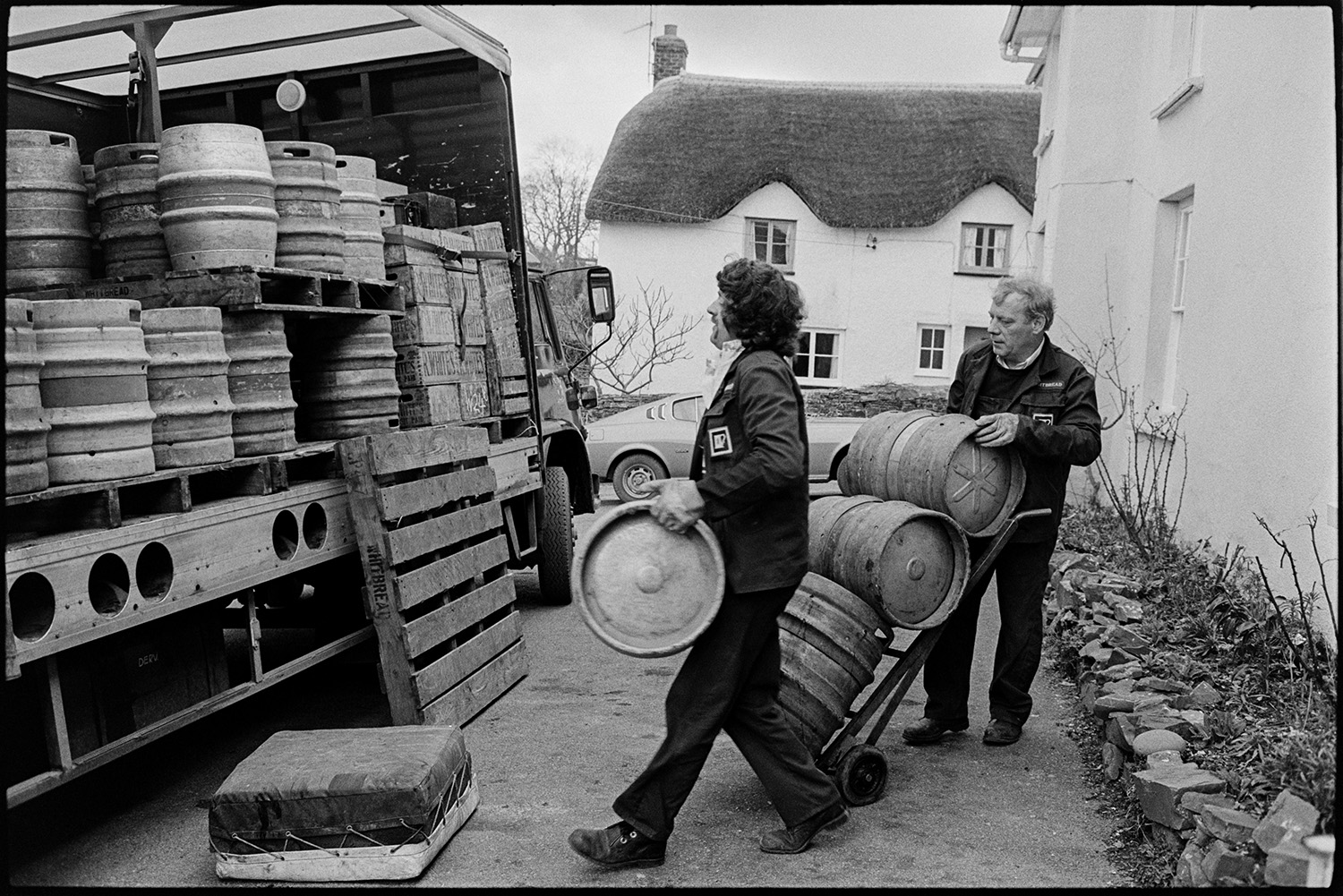 Men unloading barrels of beer from lorry outside pub. 
[Two men unloading barrels of beer from a lorry outside the Rams Head pub in Dolton. A thatched cottage with eyebrow eaves can be seen in the background.]