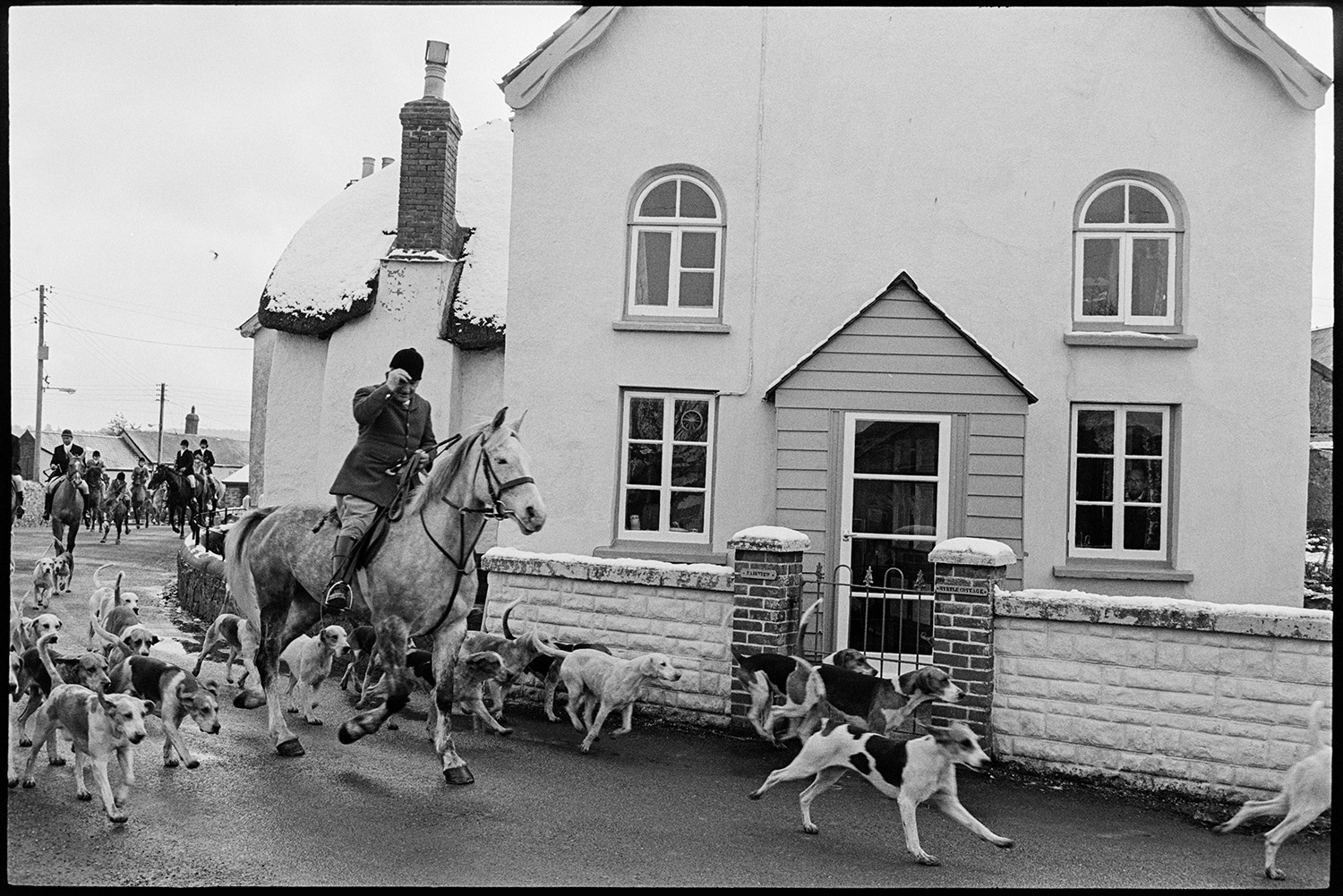 Hunt meet in village, setting off. 
[Huntsmen setting off on horseback, with hounds, through Dolton on a hunt. They are passing a house with a porch. A thatched cottage covered with snow can also be seen in the background.]